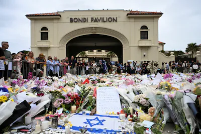 people-stand-near-flowers-laid-as-a-tribute-at-bondi-beach-to-honour-the-victims-of-a-mass-shooting.webp