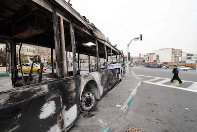 a-photograph-shows-the-wreckage-of-a-burnt-bus-bearing-a-banner-that-reads-this-was-one-of-tehrans-n.webp